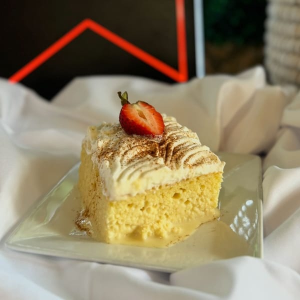 Slice of Tres Leches cake with whipped cream, cinnamon, and a strawberry on a white plate.