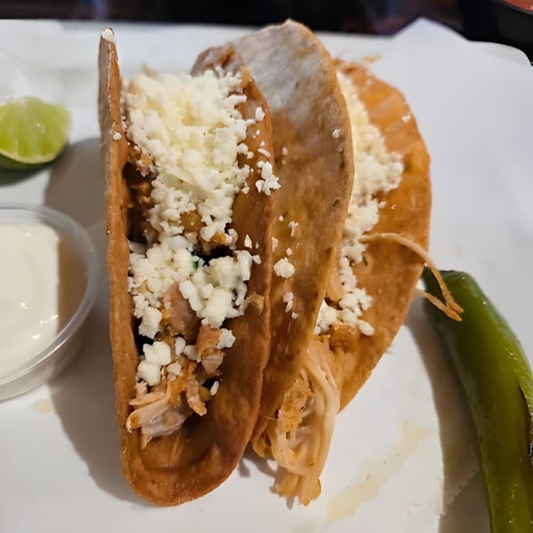 Two crispy tacos filled with shredded chicken and cheese, served with lime, sour cream, and a jalapeño.