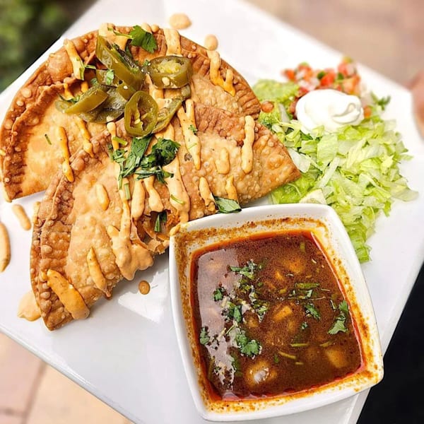 Crispy Mexican empanadas topped with jalapeños and sauce, served with a side of consommé and salad.