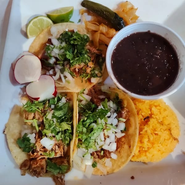 Three Mexican street tacos with carnitas, cilantro, and onions, served with rice, black beans, and lime.