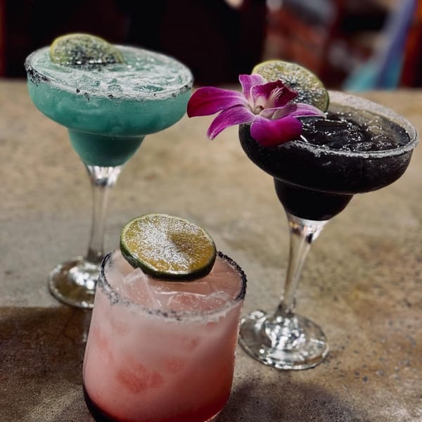 Three colorful margaritas, blue, black, and pink, garnished with lime and an orchid on a rustic table.